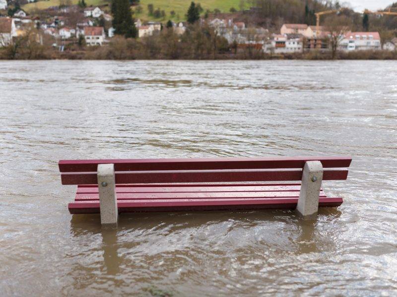 Überschwemmung an einem Fluss: Eine rote Bank ist von Wasser umspült.